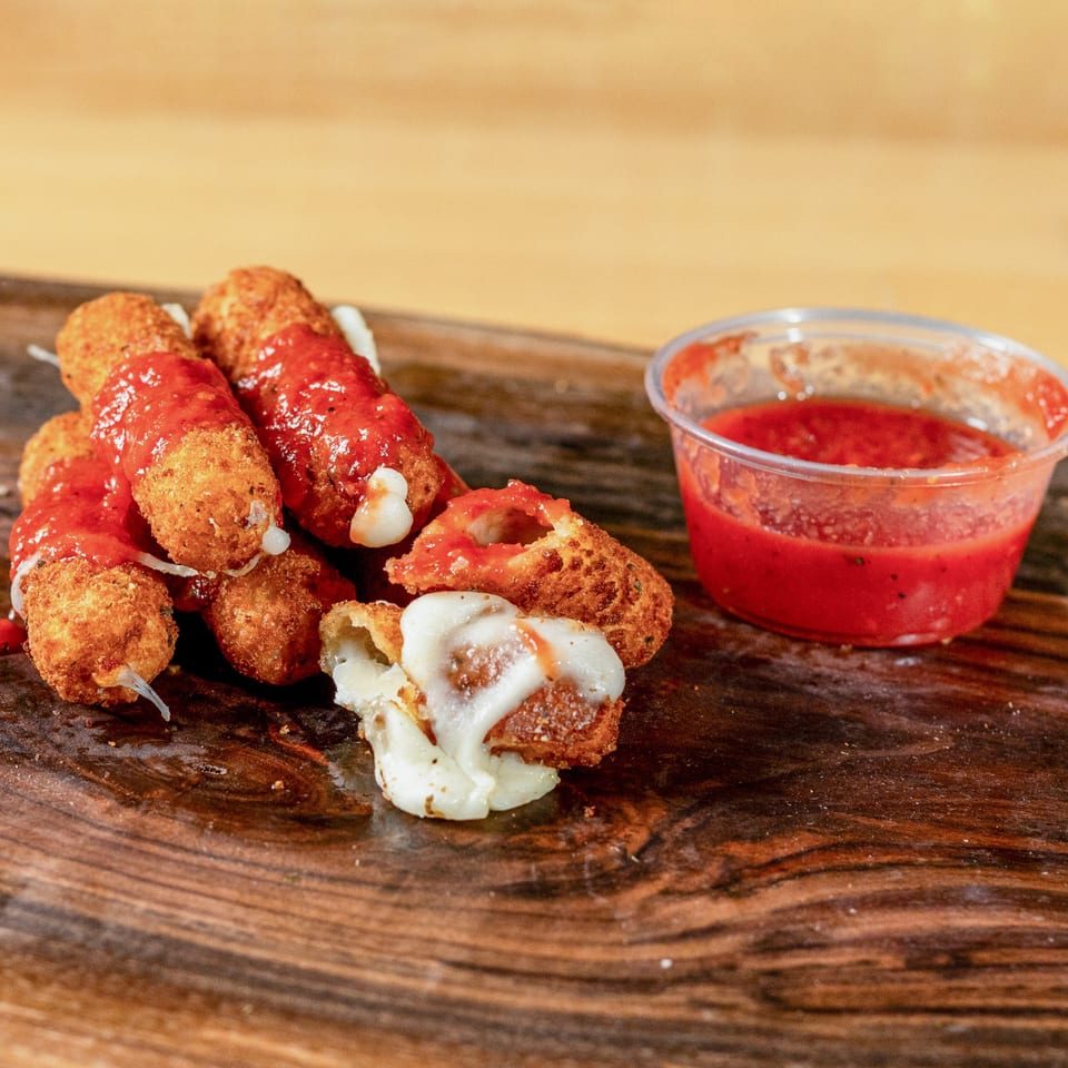 Golden-brown mozzarella sticks, some broken with melted cheese, drizzled with red sauce next to a sauce cup.