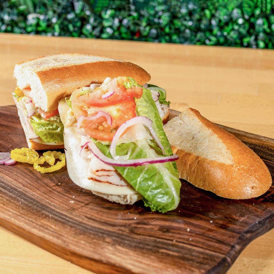 A large, fresh La Pizzeria sandwich with turkey, cheese, lettuce, tomato, and onion on a wooden board.