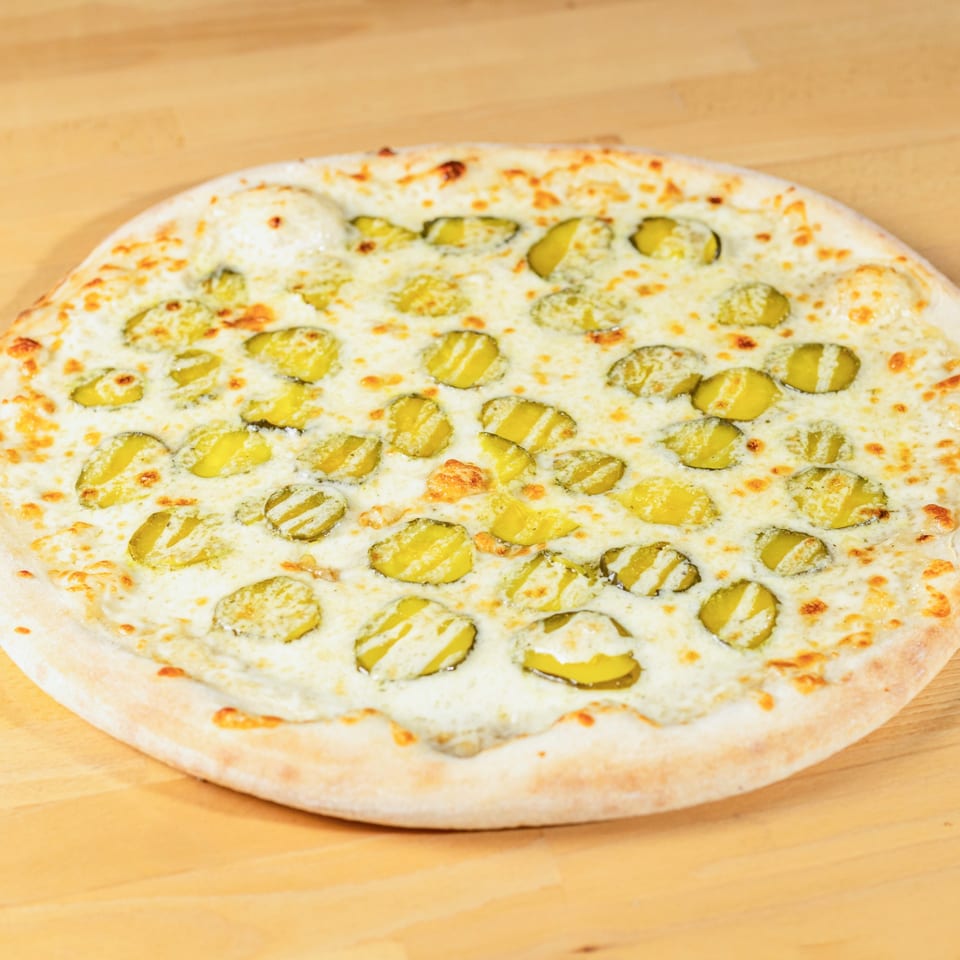 A La Pizzeria pizza with a white sauce base and sliced pickles on a wooden table.