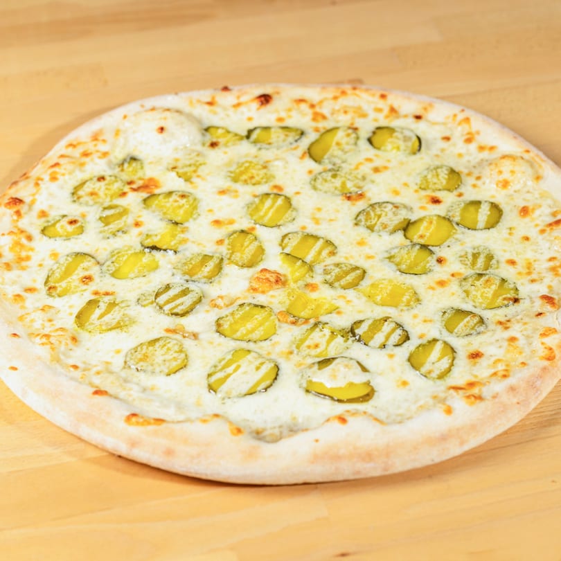 A La Pizzeria pizza with a white sauce base and sliced pickles on a wooden table.