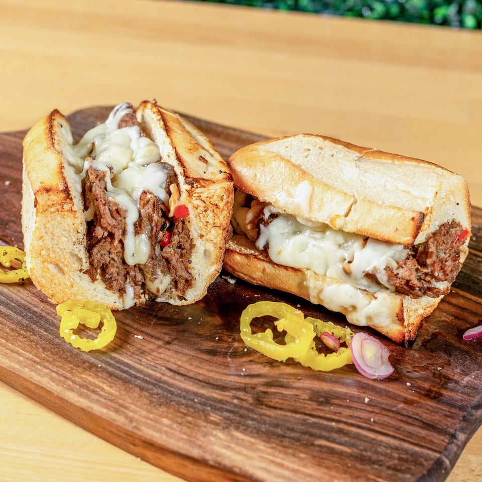 Two halves of a cheesy steak sandwich on a wooden board with peppers and onions.